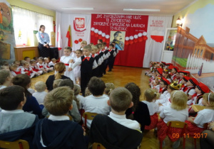 Na tle biało czerwonej dekoracji stoją w dwóch rzędach odświętnie ubrane dzieci. Pozostałe dzieci ich podziwiają.